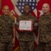 Three United States Marine Corps personnel are posing for a formal photograph. The central figure is holding a United States Marine Corps Certificate of Commendation with a red border. They are standing in front of American and Marine Corps flags in what appears to be an official office setting.