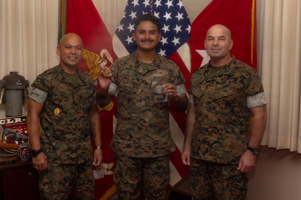 Three U.S. Marines in MARPAT camouflage uniforms pose for a formal recognition photo. The central figure is holding a clear glass award plaque while the figure on the left holds what appears to be a coin or medal. The setting appears to be an official military office with American and Marine Corps flags in the background.