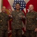 Three U.S. Marines in MARPAT camouflage uniforms pose for a formal recognition photo. The central figure is holding a clear glass award plaque while the figure on the left holds what appears to be a coin or medal. The setting appears to be an official military office with American and Marine Corps flags in the background.