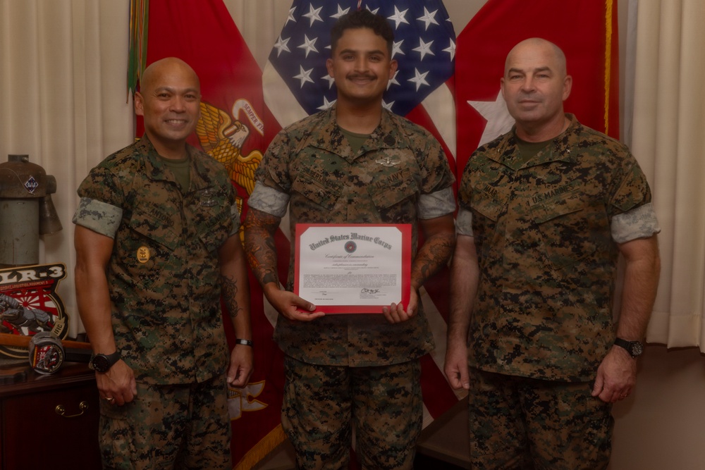 Three military personnel in USMC MARPAT camouflage uniforms pose for a formal photo. The center individual holds a United States Marine Corps Certificate of Commendation with a red border. The setting appears to be an official military office with flags and unit memorabilia visible in the background.