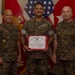 Three military personnel in USMC MARPAT camouflage uniforms pose for a formal photo. The center individual holds a United States Marine Corps Certificate of Commendation with a red border. The setting appears to be an official military office with flags and unit memorabilia visible in the background.