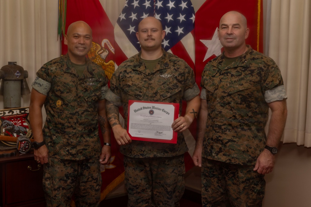 Three military personnel in USMC MARPAT camouflage uniforms pose for a formal photo. The center individual is holding a United States Marine Corps Certificate of Commendation with a red border. The setting appears to be an official military office with flags and unit insignia visible in the background.