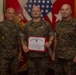 Three military personnel in USMC MARPAT camouflage uniforms pose for a formal photo. The center individual is holding a United States Marine Corps Certificate of Commendation with a red border. The setting appears to be an official military office with flags and unit insignia visible in the background.