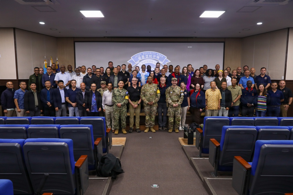 A large group photograph taken inside a conference or briefing room, with approximately 60-80 individuals posing together in front of a projection screen displaying the 'Joint Security Area' (JSA) emblem with Korean text (공동경비구역). The group includes a mix of military personnel in various uniforms and civilians in casual attire, suggesting a multinational or joint military-civilian gathering. The setting appears to be at Camp Bonifas or a JSA facility on the Korean Peninsula.