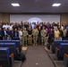 A large group photograph taken inside a conference or briefing room, with approximately 60-80 individuals posing together in front of a projection screen displaying the 'Joint Security Area' (JSA) emblem with Korean text (공동경비구역). The group includes a mix of military personnel in various uniforms and civilians in casual attire, suggesting a multinational or joint military-civilian gathering. The setting appears to be at Camp Bonifas or a JSA facility on the Korean Peninsula.