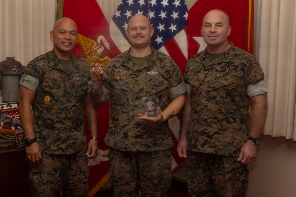 Three military personnel in MARPAT (Marine Pattern) camouflage uniforms pose for a formal recognition photo. The center individual is holding a challenge coin and a clear glass/acrylic award plaque, indicating an awards or recognition ceremony. American and Marine Corps flags are visible in the background.