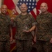 Three military personnel in MARPAT (Marine Pattern) camouflage uniforms pose for a formal recognition photo. The center individual is holding a challenge coin and a clear glass/acrylic award plaque, indicating an awards or recognition ceremony. American and Marine Corps flags are visible in the background.
