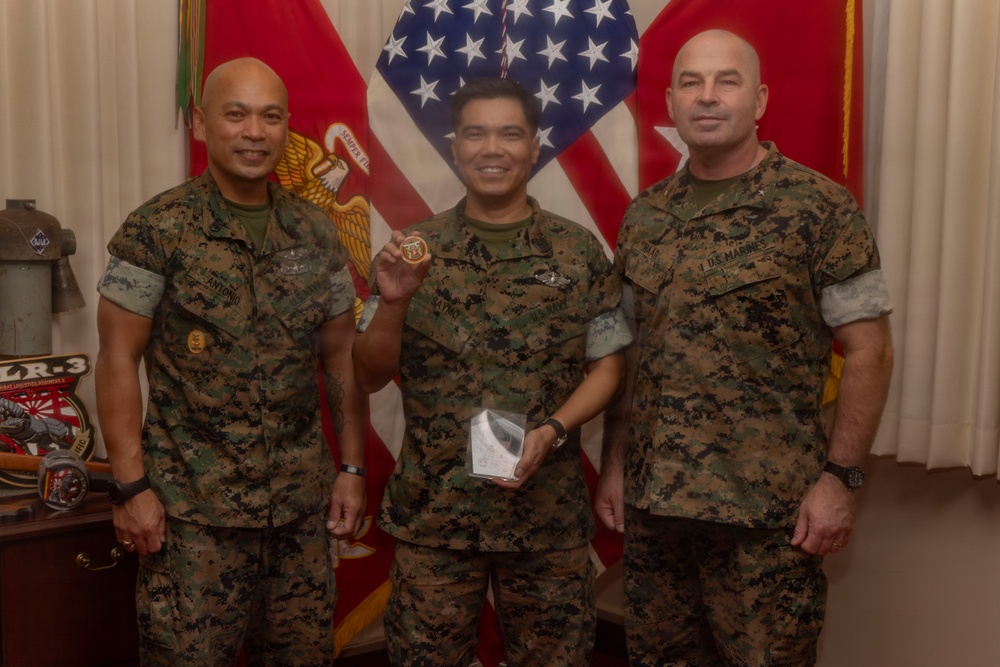 Three U.S. Marines in MARPAT camouflage uniforms pose for a formal photo, likely during a recognition or award ceremony. The central figure is holding up a challenge coin and a small gift box, suggesting he is the recipient of an honor. They are standing in front of U.S. and Marine Corps flags in what appears to be an official office or ceremonial space.