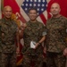 Three U.S. Marines in MARPAT camouflage uniforms pose for a formal photo, likely during a recognition or award ceremony. The central figure is holding up a challenge coin and a small gift box, suggesting he is the recipient of an honor. They are standing in front of U.S. and Marine Corps flags in what appears to be an official office or ceremonial space.