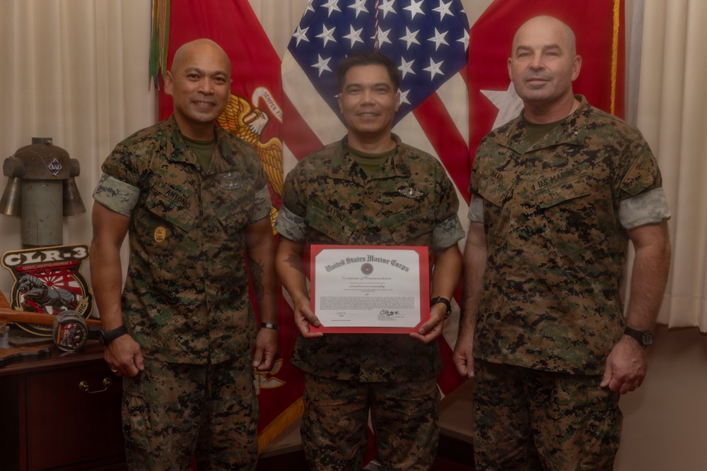 Three U.S. Marine Corps personnel in MARPAT camouflage uniforms pose for a formal photo. The central figure holds a United States Marine Corps Certificate of Commendation with a red border. The setting appears to be a military office with flags and unit insignia visible in the background.