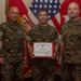 Three U.S. Marine Corps personnel in MARPAT camouflage uniforms pose for a formal photo. The central figure holds a United States Marine Corps Certificate of Commendation with a red border. The setting appears to be a military office with flags and unit insignia visible in the background.