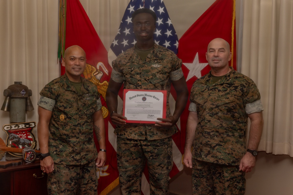Three United States Marine Corps personnel are posed for a formal recognition photo. The center Marine is holding a United States Marine Corps certificate with a red border, likely a commendation or award certificate. The setting appears to be an official military office with flags and unit insignia visible in the background.