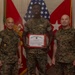 Three United States Marine Corps personnel are posed for a formal recognition photo. The center Marine is holding a United States Marine Corps certificate with a red border, likely a commendation or award certificate. The setting appears to be an official military office with flags and unit insignia visible in the background.