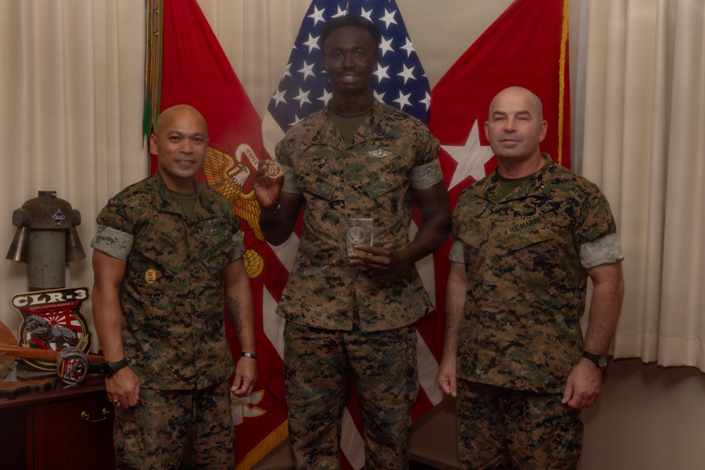 Three U.S. Marines in MARPAT camouflage uniforms pose for a formal photo in what appears to be a military office or ceremonial room. The center Marine is holding a coin and a glass award/plaque, suggesting a recognition or award ceremony. Two red general officer flags with stars are visible in the background alongside a U.S. flag.