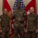 Three U.S. Marines in MARPAT camouflage uniforms pose for a formal photo in what appears to be a military office or ceremonial room. The center Marine is holding a coin and a glass award/plaque, suggesting a recognition or award ceremony. Two red general officer flags with stars are visible in the background alongside a U.S. flag.