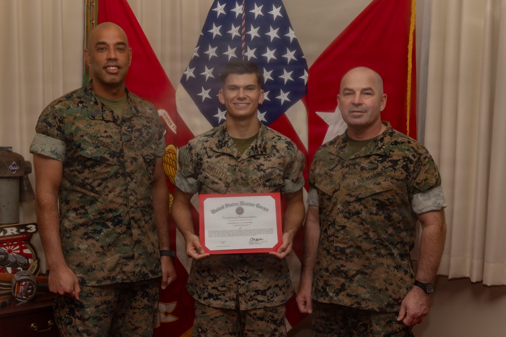 Three United States Marines in MARPAT camouflage uniforms pose for a formal photo. The center Marine is holding a United States Marine Corps Certificate of Commendation with a red border. The setting appears to be an official military office with American and Marine Corps flags in the background.