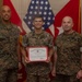 Three United States Marines in MARPAT camouflage uniforms pose for a formal photo. The center Marine is holding a United States Marine Corps Certificate of Commendation with a red border. The setting appears to be an official military office with American and Marine Corps flags in the background.