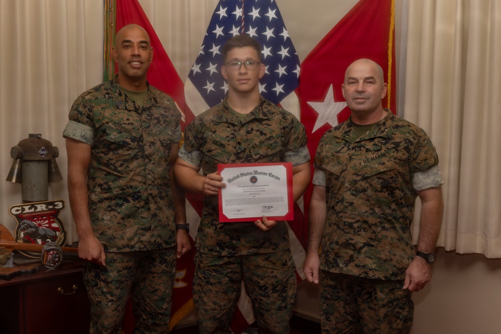 Three United States Marines in MARPAT camouflage uniforms pose for a formal photo. The central Marine is holding a United States Marine Corps Certificate of Commendation with a red border. The setting appears to be a military office with flags and unit insignia visible in the background.