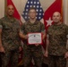 Three United States Marines in MARPAT camouflage uniforms pose for a formal photo. The central Marine is holding a United States Marine Corps Certificate of Commendation with a red border. The setting appears to be a military office with flags and unit insignia visible in the background.