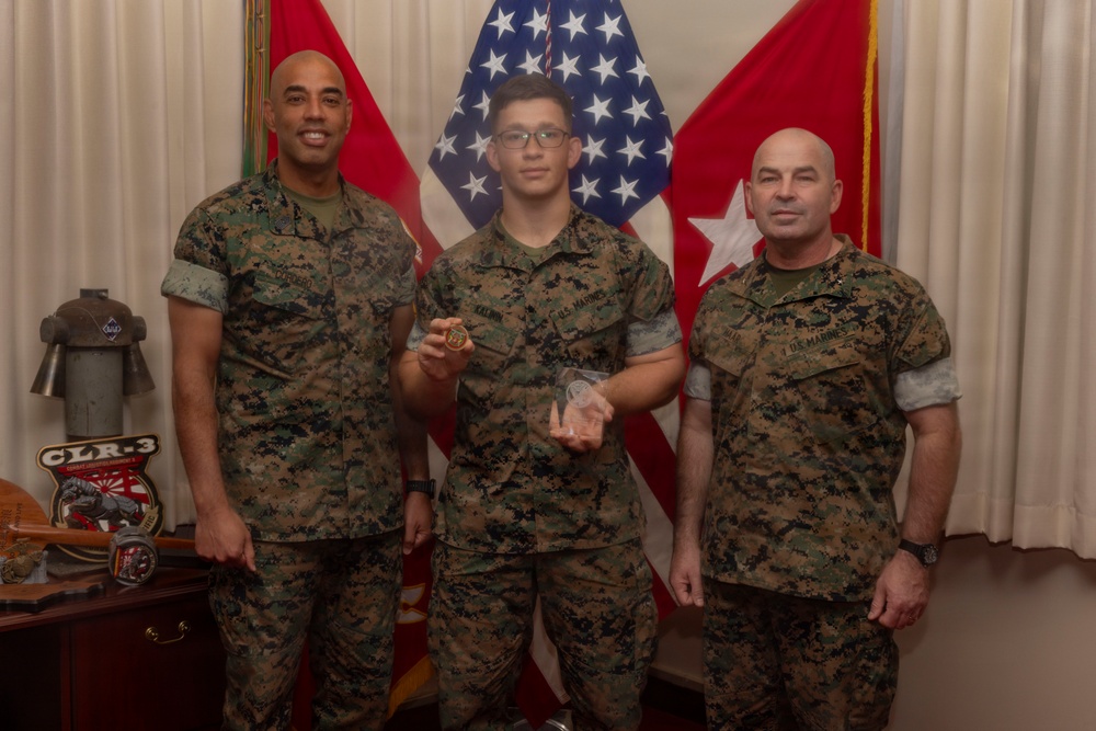 Three U.S. Marines in MARPAT camouflage uniforms pose for a formal photo in what appears to be a command office or ceremonial space. The center Marine is holding what appears to be a challenge coin and a clear acrylic award, suggesting a recognition or award ceremony. Red and white military flags and a U.S. flag are displayed in the background.