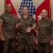 Three U.S. Marines in MARPAT camouflage uniforms pose for a formal photo in what appears to be a command office or ceremonial space. The center Marine is holding what appears to be a challenge coin and a clear acrylic award, suggesting a recognition or award ceremony. Red and white military flags and a U.S. flag are displayed in the background.