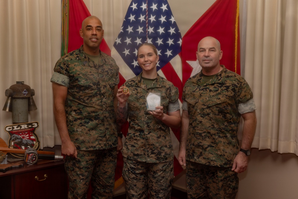 Three U.S. Marines in digital camouflage (MARPAT) uniforms pose for a formal recognition photo. The central figure, a female Marine, is holding what appears to be a challenge coin and a clear acrylic award, suggesting an award or commendation ceremony. They are standing in front of an American flag and red military unit flags.