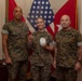 Three U.S. Marines in digital camouflage (MARPAT) uniforms pose for a formal recognition photo. The central figure, a female Marine, is holding what appears to be a challenge coin and a clear acrylic award, suggesting an award or commendation ceremony. They are standing in front of an American flag and red military unit flags.