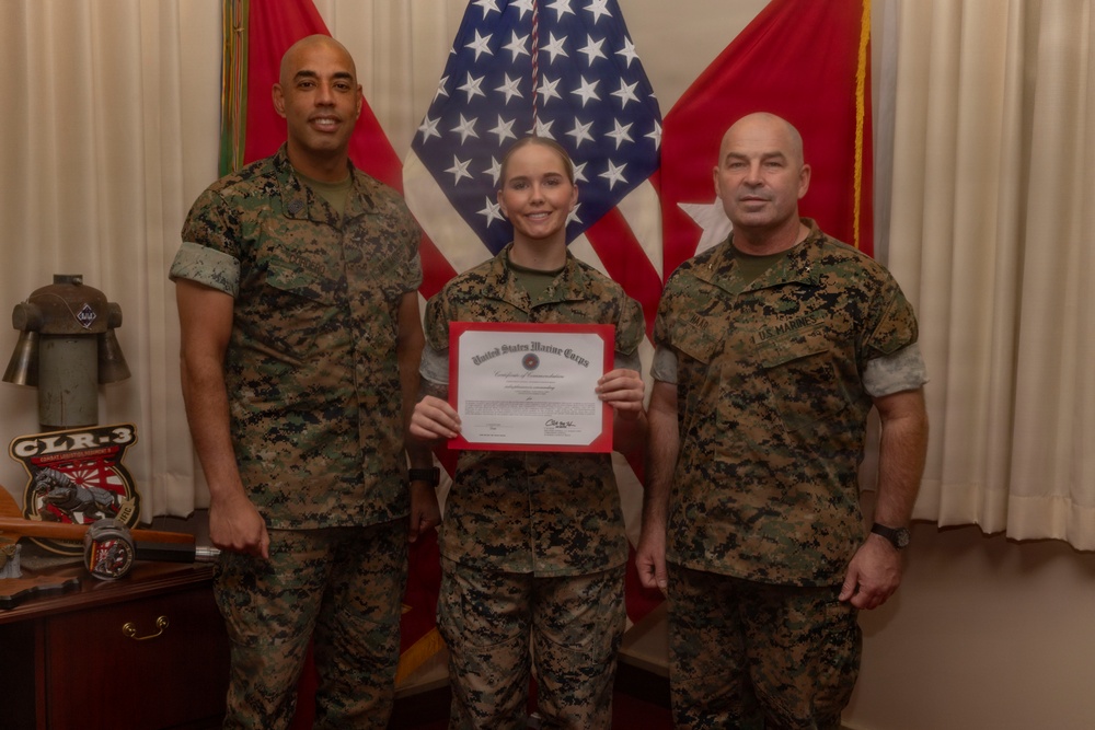 Three United States Marine Corps personnel in MARPAT camouflage uniforms pose for a formal photo. The central figure, a female Marine, holds a United States Marine Corps Certificate of Commendation with a red border. The setting appears to be an official military office with flags and unit insignia visible in the background.