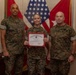 Three United States Marine Corps personnel in MARPAT camouflage uniforms pose for a formal photo. The central figure, a female Marine, holds a United States Marine Corps Certificate of Commendation with a red border. The setting appears to be an official military office with flags and unit insignia visible in the background.