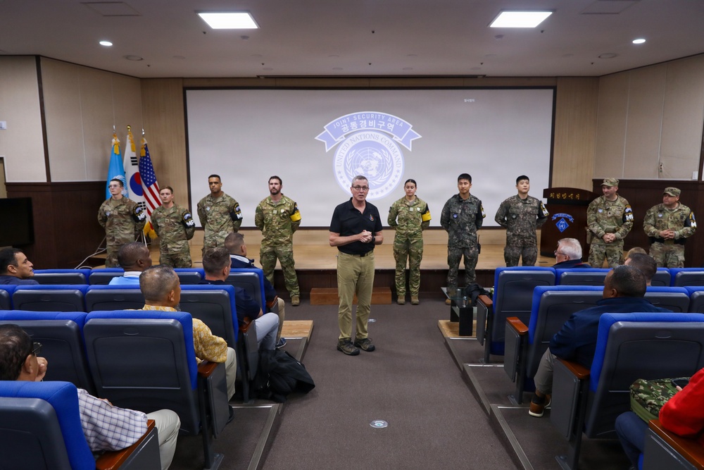 A briefing or orientation session is taking place in an auditorium at the Joint Security Area (JSA), United Nations Command, likely at Camp Bonifas near the Korean DMZ. A civilian presenter stands at the front addressing a seated audience, while a mixed group of military personnel in US Army multicam uniforms and Republic of Korea Army uniforms stand in a line behind him. The JSA/United Nations Command logo is projected on the screen behind the group.