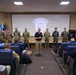 A briefing or orientation session is taking place in an auditorium at the Joint Security Area (JSA), United Nations Command, likely at Camp Bonifas near the Korean DMZ. A civilian presenter stands at the front addressing a seated audience, while a mixed group of military personnel in US Army multicam uniforms and Republic of Korea Army uniforms stand in a line behind him. The JSA/United Nations Command logo is projected on the screen behind the group.