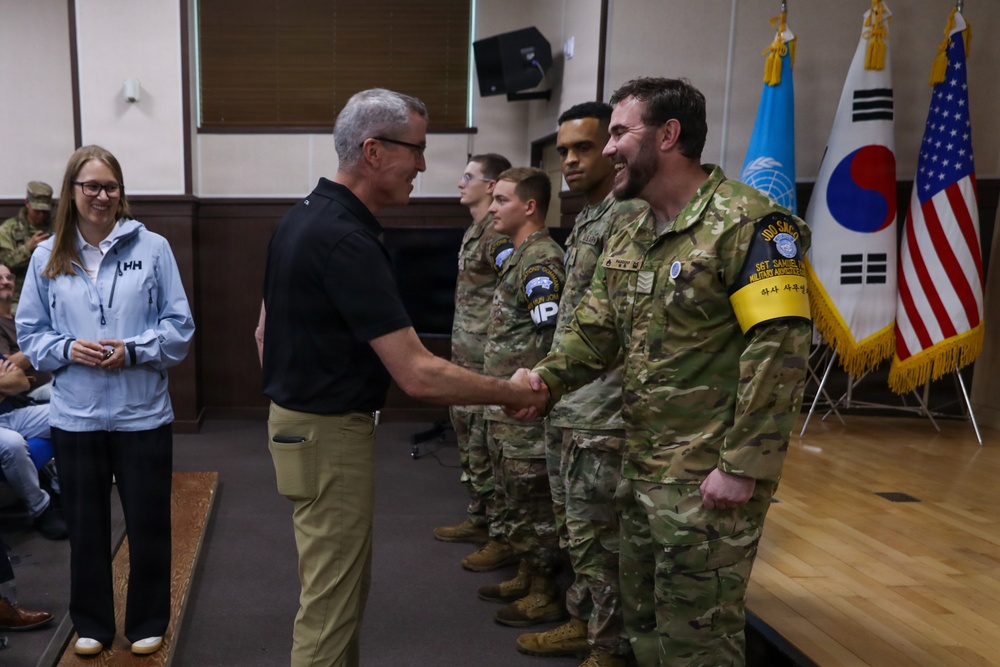 A civilian man in a black polo shirt and khaki pants is shaking hands with a uniformed soldier wearing a yellow armband reading 'Military Armistice' in a formal indoor setting. Several other soldiers in multicam uniforms are lined up in the background, with flags of the United Nations, South Korea, and the United States visible. A woman in a light blue Helly Hansen jacket observes and smiles in the background.
