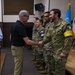 A civilian man in a black polo shirt and khaki pants is shaking hands with a uniformed soldier wearing a yellow armband reading 'Military Armistice' in a formal indoor setting. Several other soldiers in multicam uniforms are lined up in the background, with flags of the United Nations, South Korea, and the United States visible. A woman in a light blue Helly Hansen jacket observes and smiles in the background.