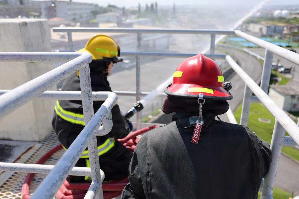 Two firefighters are positioned on an elevated platform or structure, operating a fire hose that is actively discharging water. One firefighter wears a yellow helmet with reflective striping and standard turnout gear, while the other wears a red helmet with a visible tag reading 'MIYAGI Y'. The setting appears to be an industrial or urban area, suggesting a training exercise or active firefighting operation.