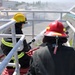 Two firefighters are positioned on an elevated platform or structure, operating a fire hose that is actively discharging water. One firefighter wears a yellow helmet with reflective striping and standard turnout gear, while the other wears a red helmet with a visible tag reading 'MIYAGI Y'. The setting appears to be an industrial or urban area, suggesting a training exercise or active firefighting operation.