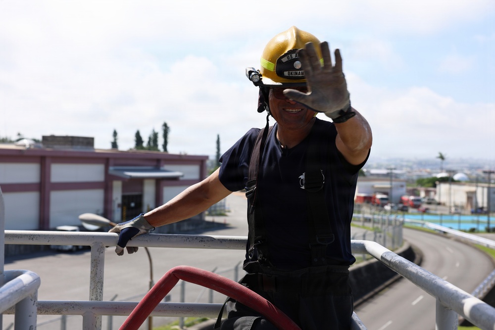 A firefighter wearing a yellow helmet labeled 'US A... OKINAWA' is positioned on an elevated platform or rooftop, raising one gloved hand toward the camera. The person is dressed in dark navy firefighting attire with suspenders and is holding or managing a red fire hose. The background reveals a military or industrial base environment consistent with Okinawa, Japan.