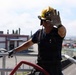 A firefighter wearing a yellow helmet labeled 'US A... OKINAWA' is positioned on an elevated platform or rooftop, raising one gloved hand toward the camera. The person is dressed in dark navy firefighting attire with suspenders and is holding or managing a red fire hose. The background reveals a military or industrial base environment consistent with Okinawa, Japan.