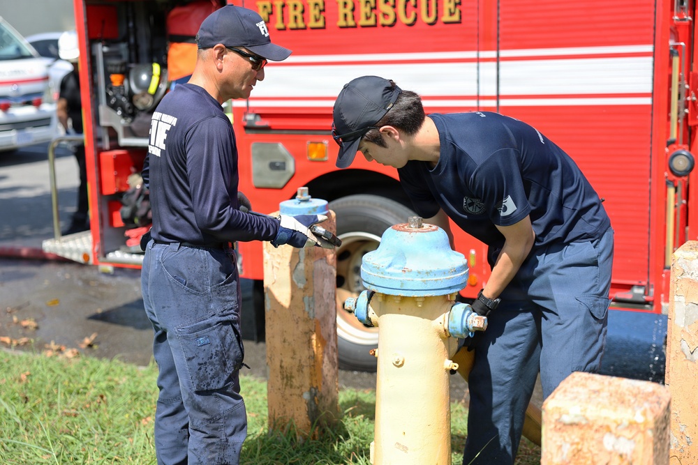 Two firefighters in navy blue uniforms are working on a fire hydrant in front of a red Fire Rescue truck. One firefighter is observing and holding a tool while the other is actively working on the hydrant's valve or cap. The scene appears to be a routine hydrant inspection or maintenance operation.