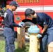 Two firefighters in navy blue uniforms are working on a fire hydrant in front of a red Fire Rescue truck. One firefighter is observing and holding a tool while the other is actively working on the hydrant's valve or cap. The scene appears to be a routine hydrant inspection or maintenance operation.