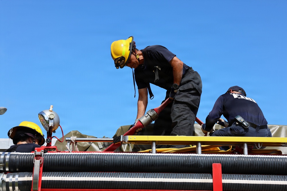 Three firefighters are working on top of a fire truck, handling hoses and equipment under a clear blue sky. The central figure, wearing a yellow helmet and black uniform, is bent over manipulating a red hose with a metal coupling. The scene appears to be either a training exercise or active emergency response preparation.