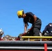 Three firefighters are working on top of a fire truck, handling hoses and equipment under a clear blue sky. The central figure, wearing a yellow helmet and black uniform, is bent over manipulating a red hose with a metal coupling. The scene appears to be either a training exercise or active emergency response preparation.