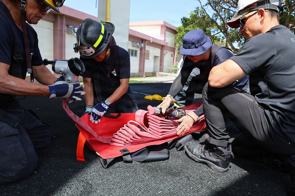 Four individuals, including at least one person wearing a firefighting helmet labeled 'ARMY', are working together to fold or pack a red fire hose on a paved outdoor surface. The group appears to be conducting equipment maintenance or training, carefully arranging the hose in accordion-style folds. The setting appears to be near a military or institutional building in a tropical environment.