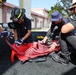 Four individuals, including at least one person wearing a firefighting helmet labeled 'ARMY', are working together to fold or pack a red fire hose on a paved outdoor surface. The group appears to be conducting equipment maintenance or training, carefully arranging the hose in accordion-style folds. The setting appears to be near a military or institutional building in a tropical environment.