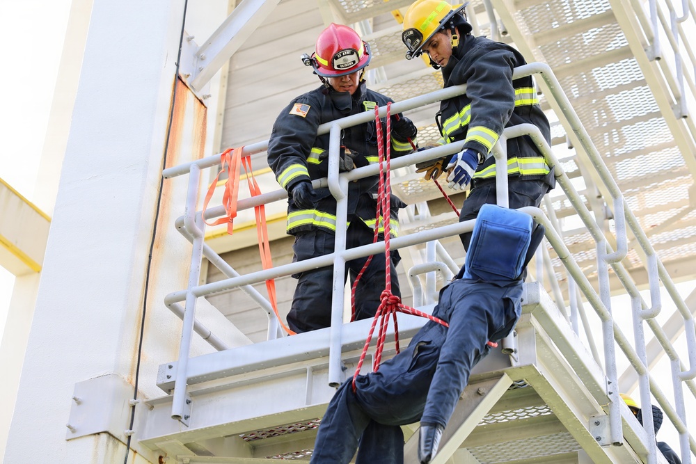 Two firefighters in full turnout gear are conducting a rope rescue training exercise on an elevated metal platform structure. They are lowering or controlling a person in blue clothing using a red rope system tied in a harness configuration. The scene appears to be a controlled training environment at a fire training tower.