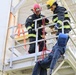 Two firefighters in full turnout gear are conducting a rope rescue training exercise on an elevated metal platform structure. They are lowering or controlling a person in blue clothing using a red rope system tied in a harness configuration. The scene appears to be a controlled training environment at a fire training tower.