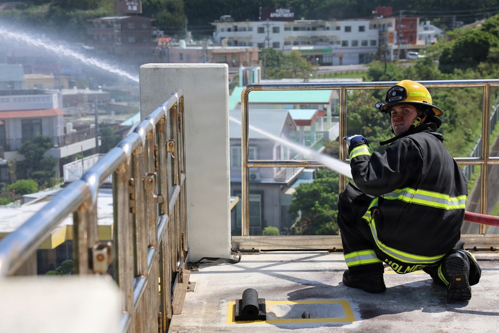 A firefighter identified as 'Holmes' is kneeling on a rooftop or elevated platform, directing a high-pressure water stream from a hose. The firefighter is wearing full turnout gear including a yellow helmet, black jacket with reflective yellow stripes, and blue gloves. The setting appears to be in a Japanese urban area, visible from signage and architecture in the background.