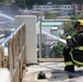 A firefighter identified as 'Holmes' is kneeling on a rooftop or elevated platform, directing a high-pressure water stream from a hose. The firefighter is wearing full turnout gear including a yellow helmet, black jacket with reflective yellow stripes, and blue gloves. The setting appears to be in a Japanese urban area, visible from signage and architecture in the background.