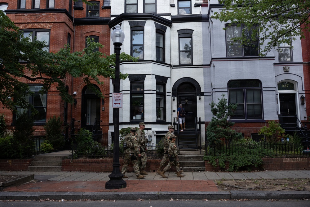 A group of military personnel in camouflage uniforms are walking along a sidewalk in front of a row of historic brick townhouses. The architecture suggests an urban neighborhood, likely in Washington D.C., based on the style of the rowhouses and a visible parking sign. A civilian can be seen entering one of the townhouses in the background.