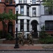 A group of military personnel in camouflage uniforms are walking along a sidewalk in front of a row of historic brick townhouses. The architecture suggests an urban neighborhood, likely in Washington D.C., based on the style of the rowhouses and a visible parking sign. A civilian can be seen entering one of the townhouses in the background.