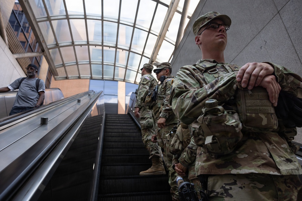 Multiple soldiers in U.S. Army camouflage (OCP/MultiCam) uniforms are riding an escalator in what appears to be a large public transit station or building with a glass-paneled ceiling. The soldiers are in full combat gear including body armor, tactical pouches, and appear to be carrying weapons. A civilian in casual clothing is visible on a parallel escalator to the left.